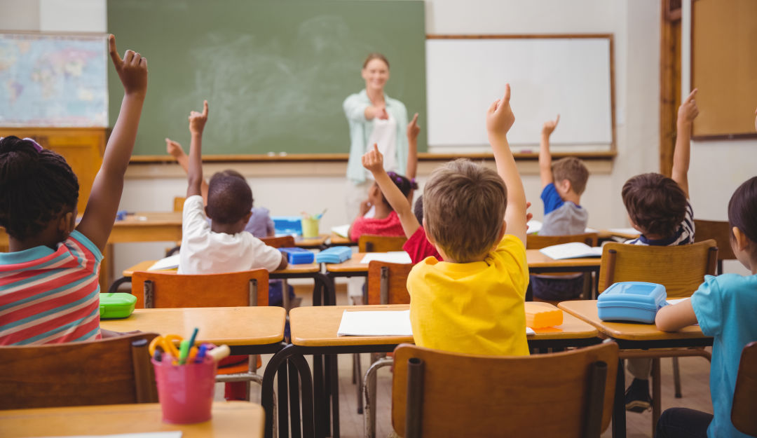 children seated at desks in a classroom and raising their hands