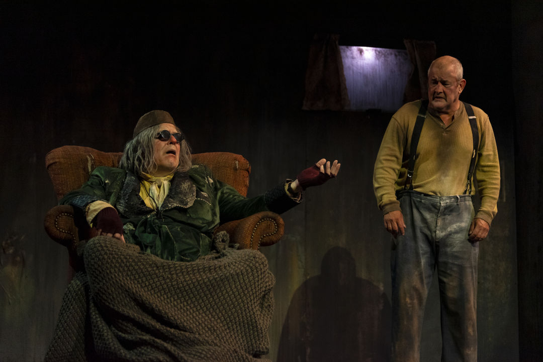 A man in a smoking jacket, glasses, and a round cap is seated next to a bald standing man in a simple shirt and pants.