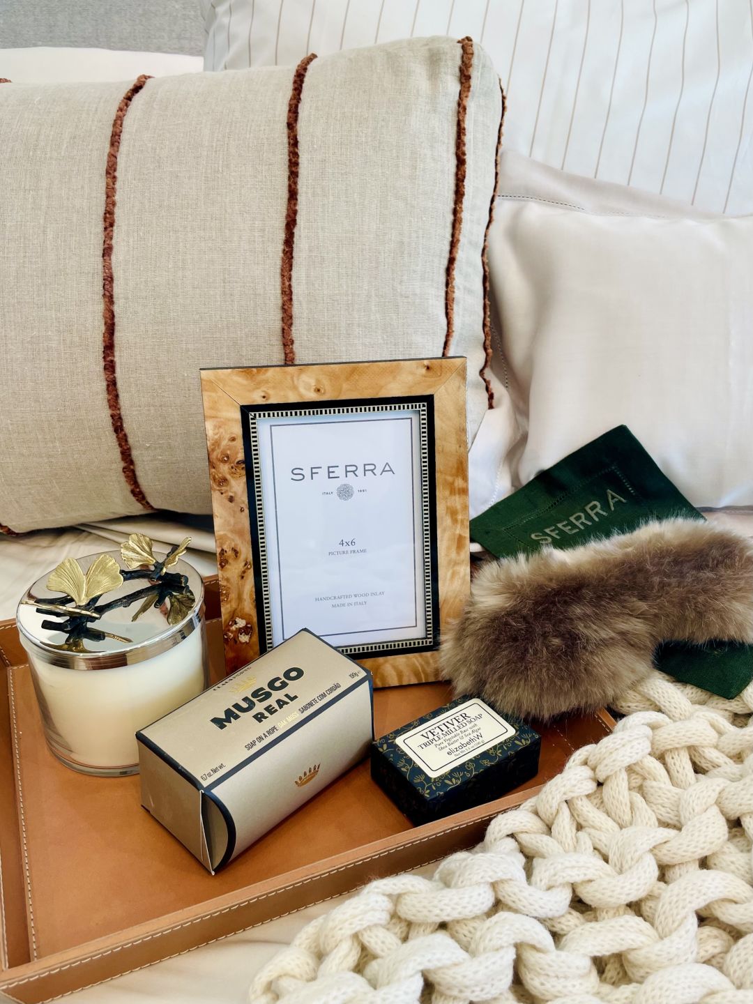 A striped throw pillow, knit throw and faux fur eye mask on a leather tray.