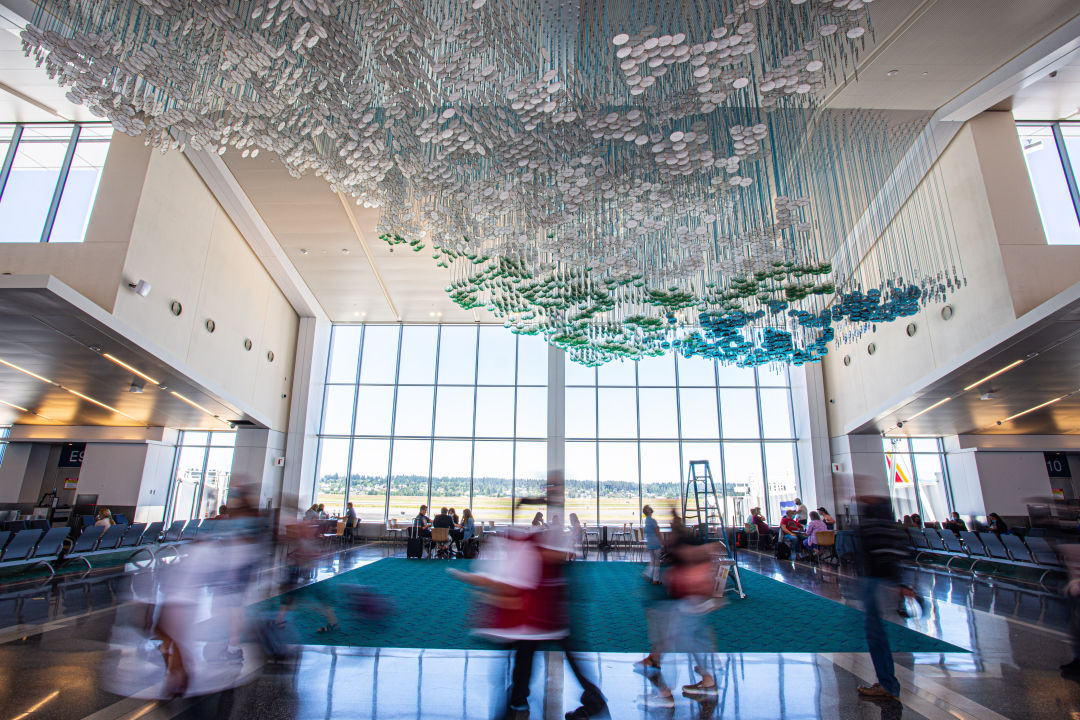 A dense groups of clear and blue discs hanging from the ceiling in front of a large window, with blurred images of people crossing in the foreground