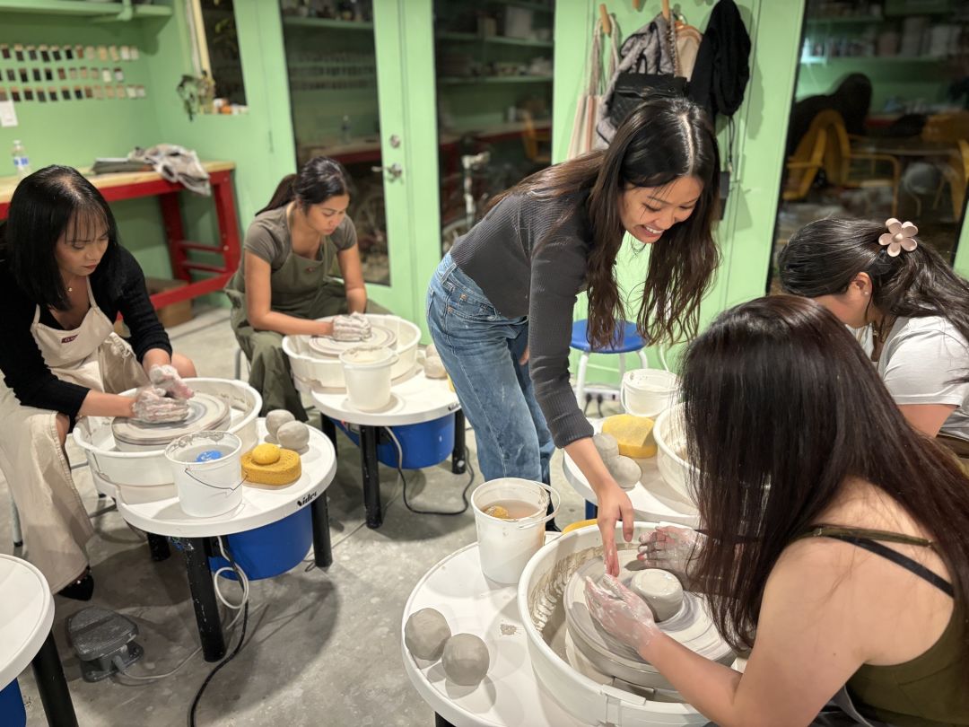 A class full of students are being instructed on pottery by a smiling teacher. They are sitting in front of pottery wheels and working with wet clay.