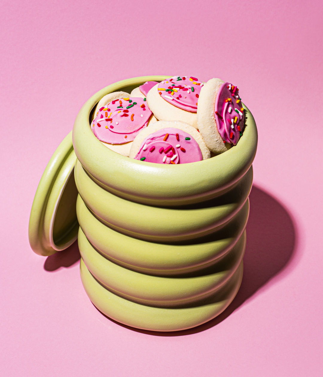 A wavy ceramic cylinder cookie jar filled with pink-frosted cookies with rainbow sprinkles