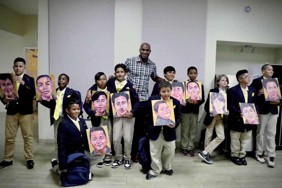 Art teacher poses with the graduating class of Visible Men Academy, holding painted portraits he gifted them.