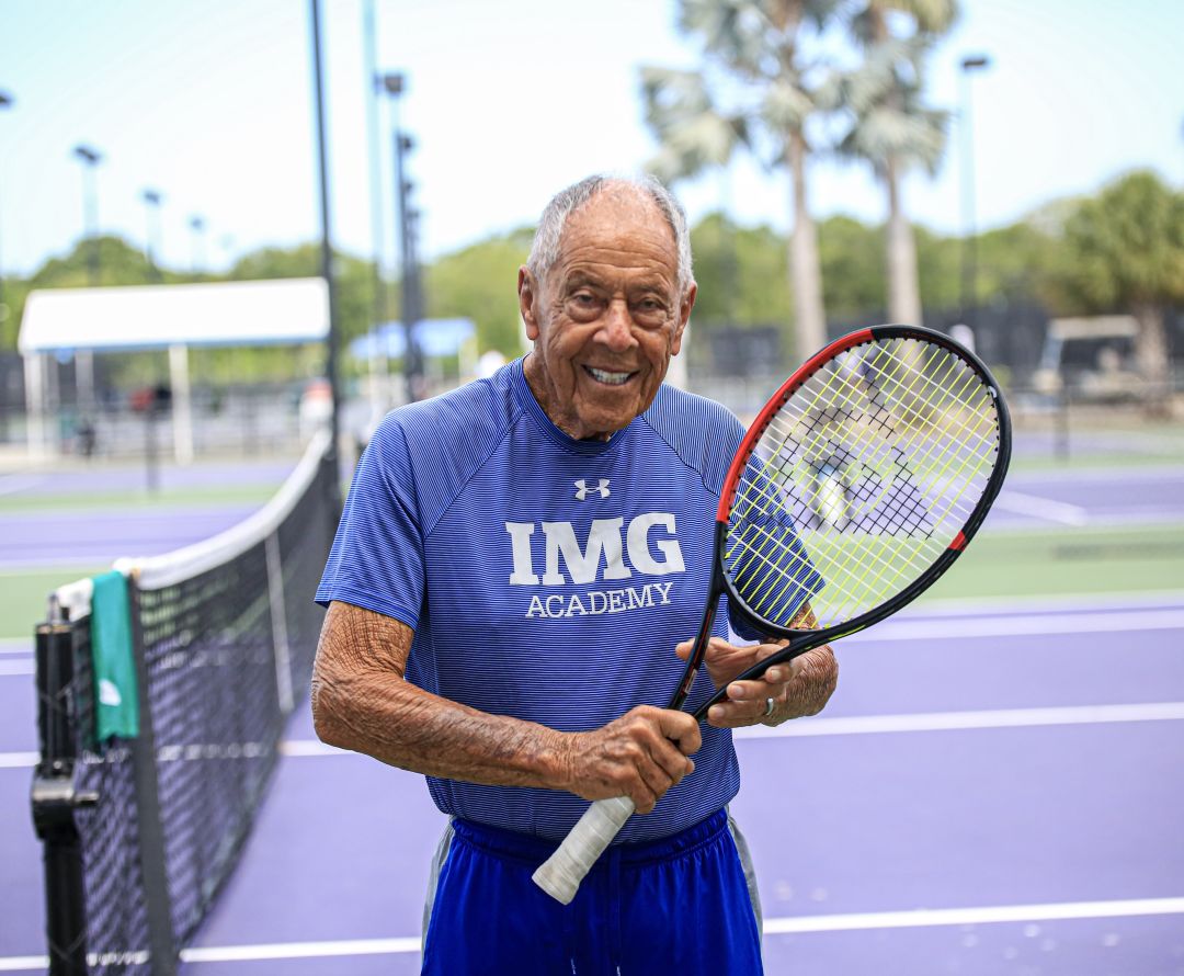 Nick Bollettieri on the tennis court wearing an IMG shirt and holdlig a tennis racket.