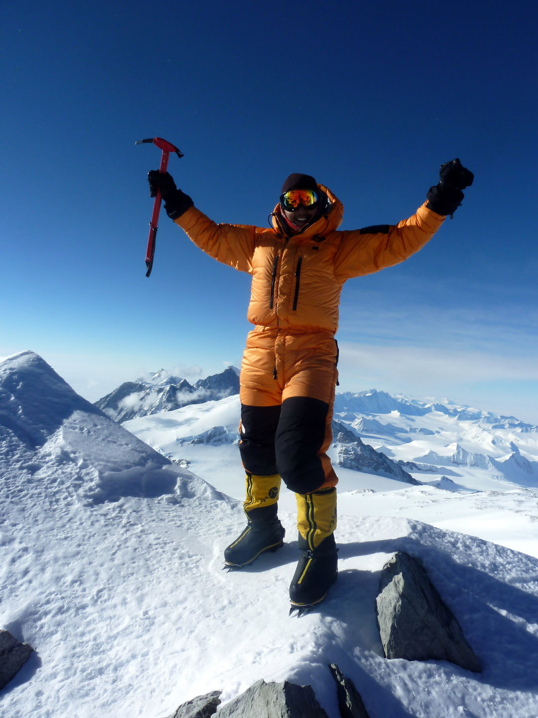 Climber in orange jacket stands on the top of a snowy mountain.