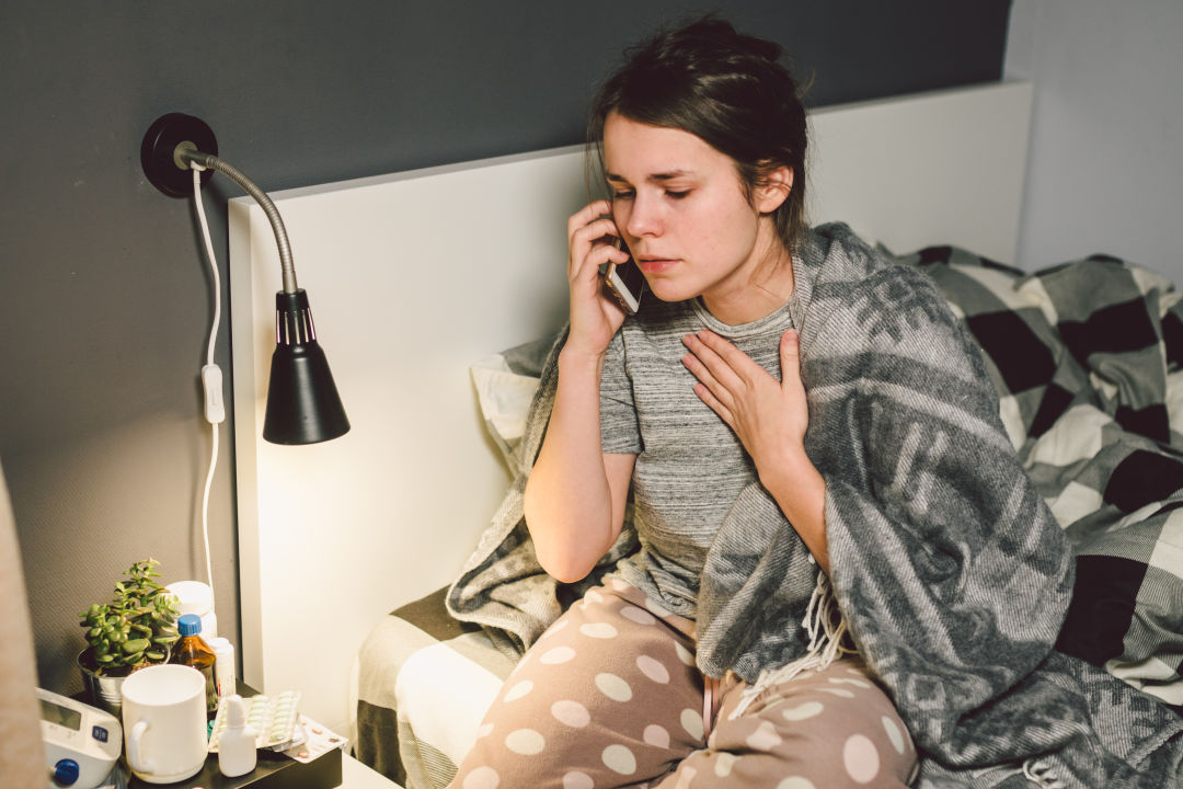 Woman sitting on bed looking concerned holding phone to ear with hand on chest, with medicine and mug on bedside table