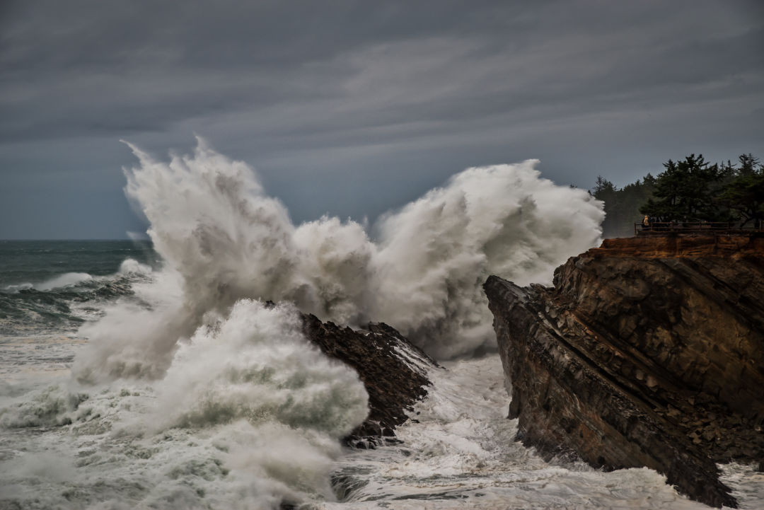 A very large crashing ocean wave on a steep rocky shore under a gray sky