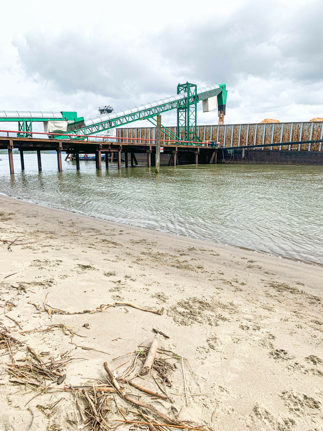 industrial conveyor dumping what looks like sand onto a barge in the river, with a sandy beach in view