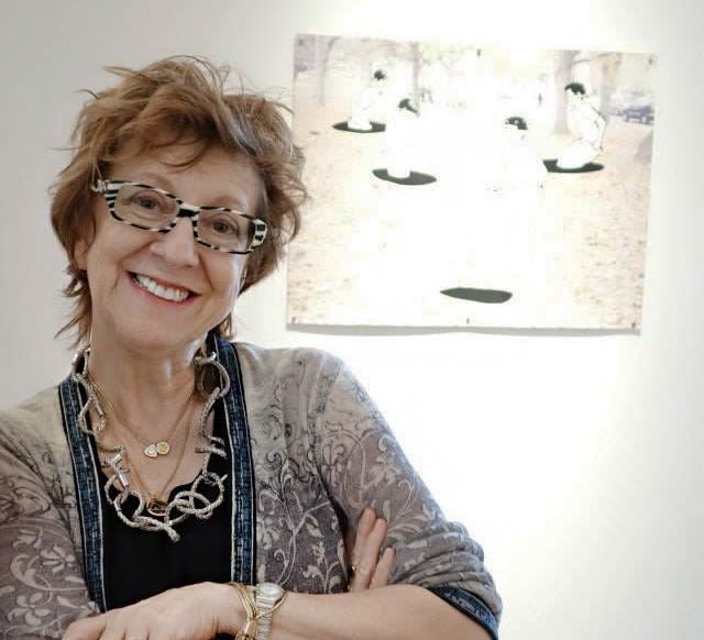 A white woman in glasses with red-grey hair and striped glasses stands next to a piece of modern art with a white background and black shapes.