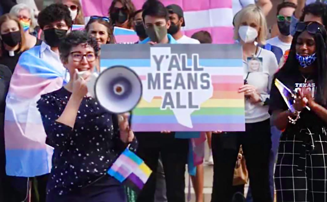 Protestors stand in front of a trans flag and a sign with a drawing of Texas on it reading "Y'all Means All." One of them speaks in a microphone.