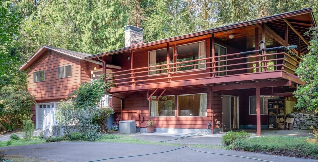 Two story red wooden home with driveway and garage, and long deck under roofline on second floor