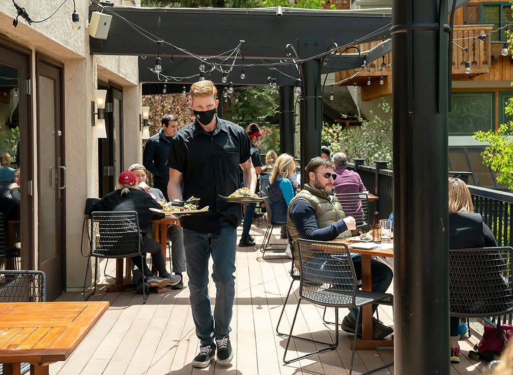 People dine at an outdoor patio while a server wearing a protective mask carries food