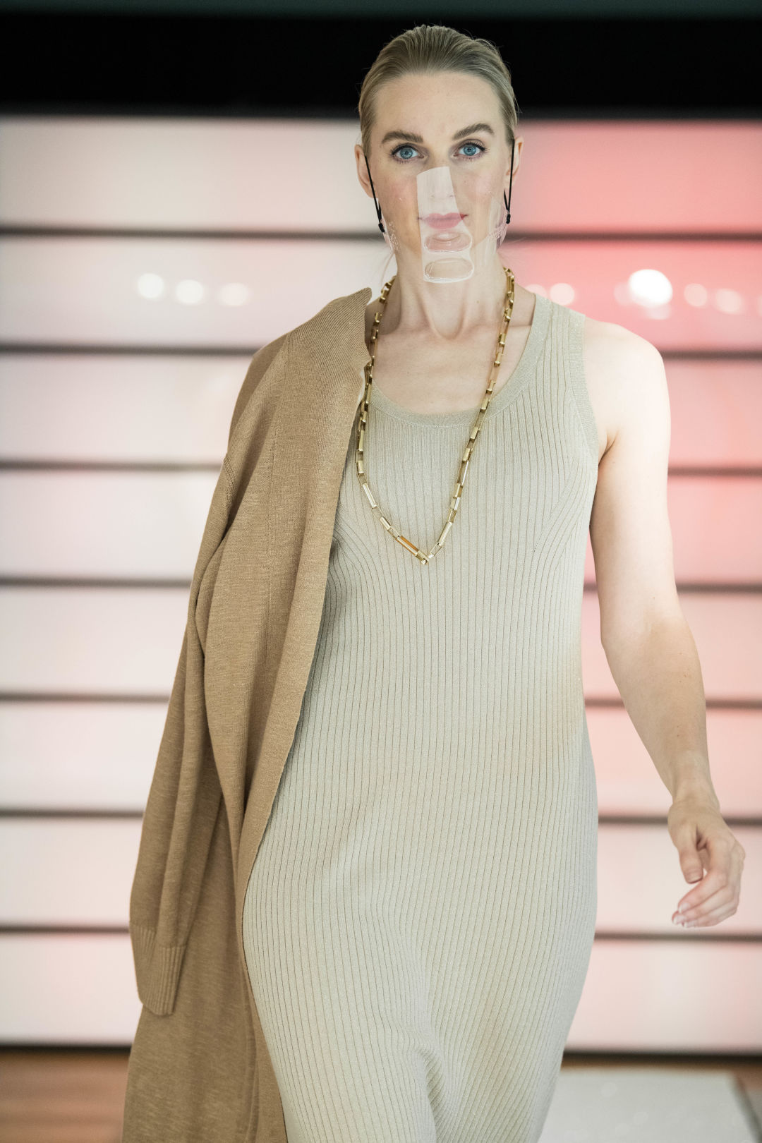 A woman walks a fashion runway in a cream colored dress with a tan sweater draped over one shoulder.
