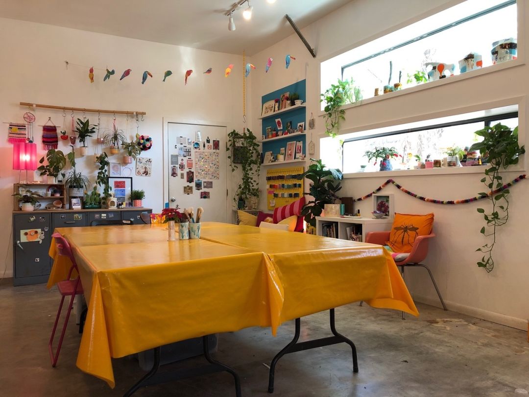 Tables covered in shiny yellow-orange tablecloths in a studio full of colorful art pieces. There's also an orange chair.