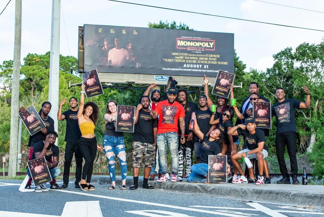 Cast and crew of the film Monopoly Money posing in front of a local billboard of the film.