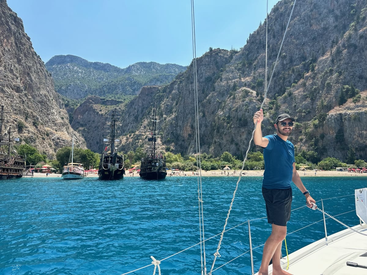 Sailor standing on bow of sailboat in clear blue water with steep cliffs behind.