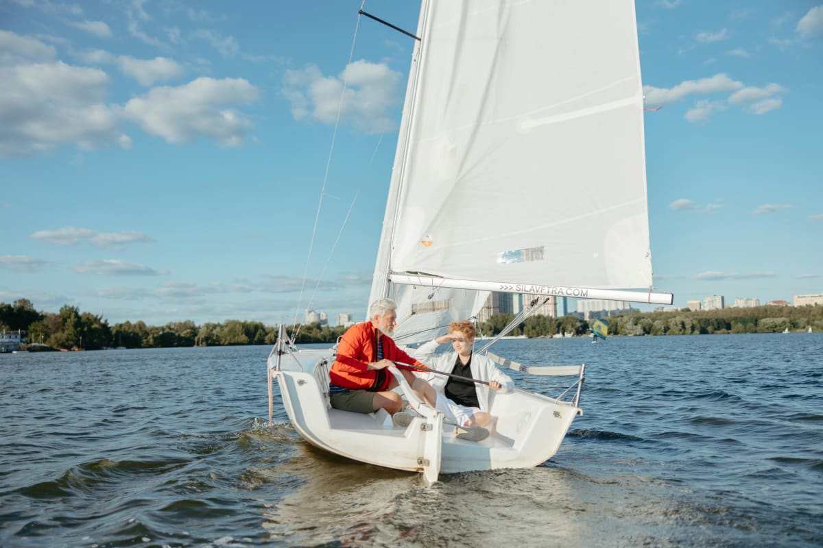 Two people sailing a small boat on a lake, with one steering and the other looking ahead in the sun.