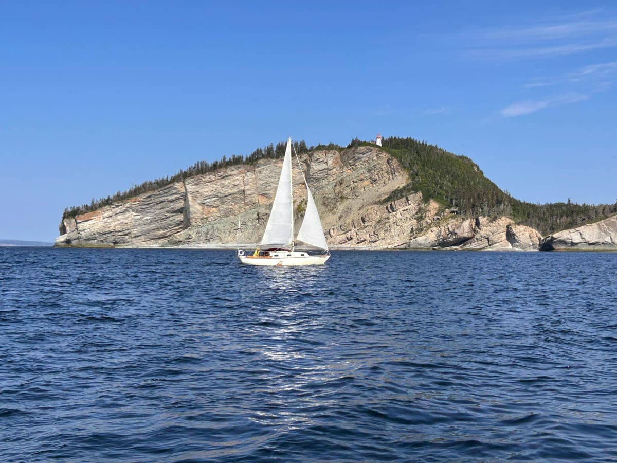 Sailboat cruising on open water near a rocky coastline with cliffs and a lighthouse in the background.