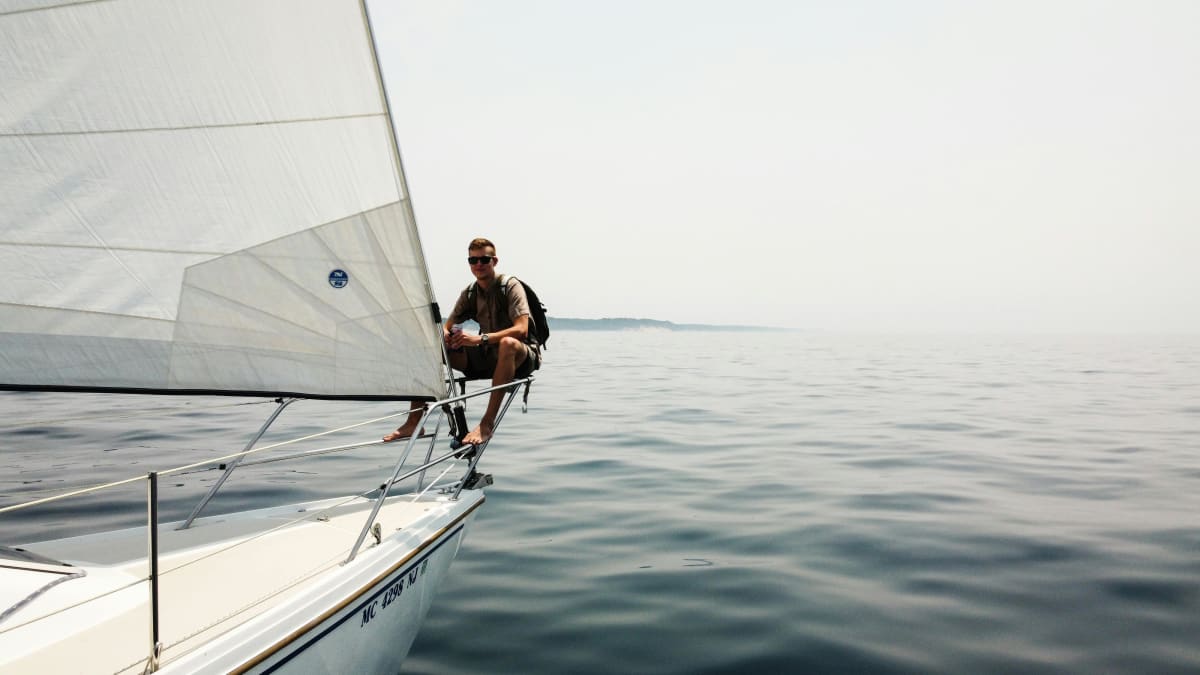 Person sitting on the bow of a sailboat with calm water and a hazy horizon in the background.