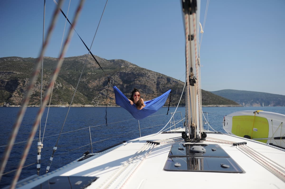 Person relaxing in a blue hammock suspended on the foredeck of a sailboat with rocky hills in the background.