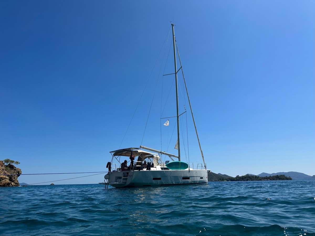 A sailboat anchored in calm blue water under a clear sky with rocky coastline in the distance.