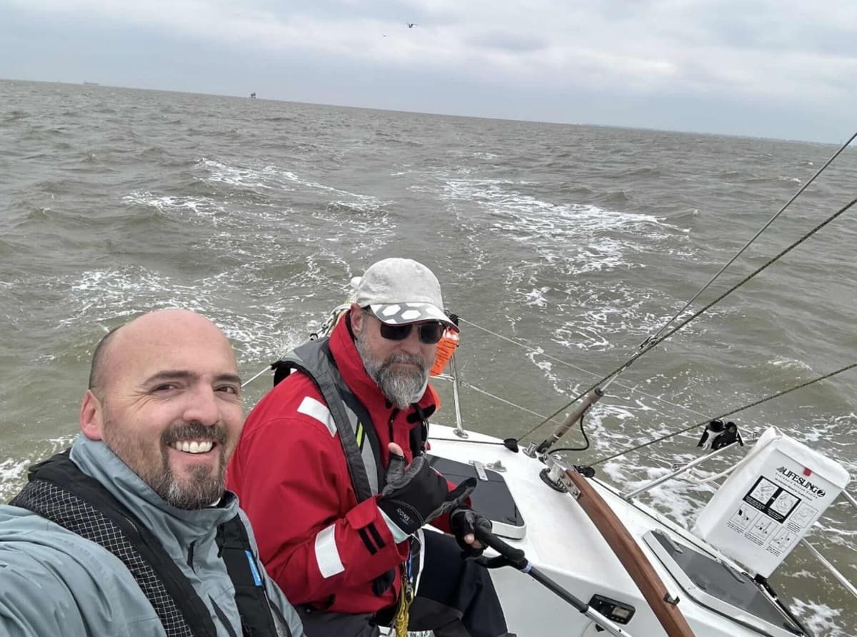 Two sailors at the helm of a sailboat navigating choppy offshore conditions.