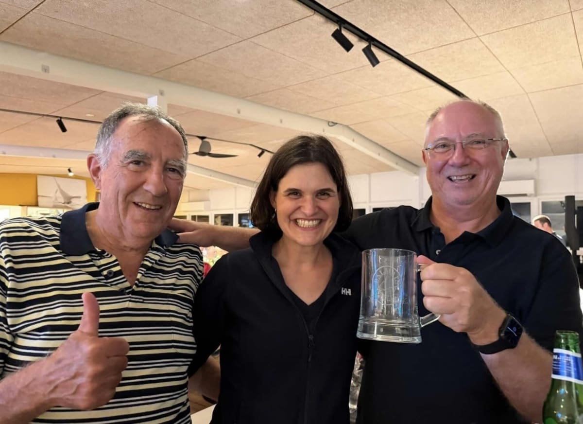 Three sailors celebrating together with smiles and drinks inside a sailing club.