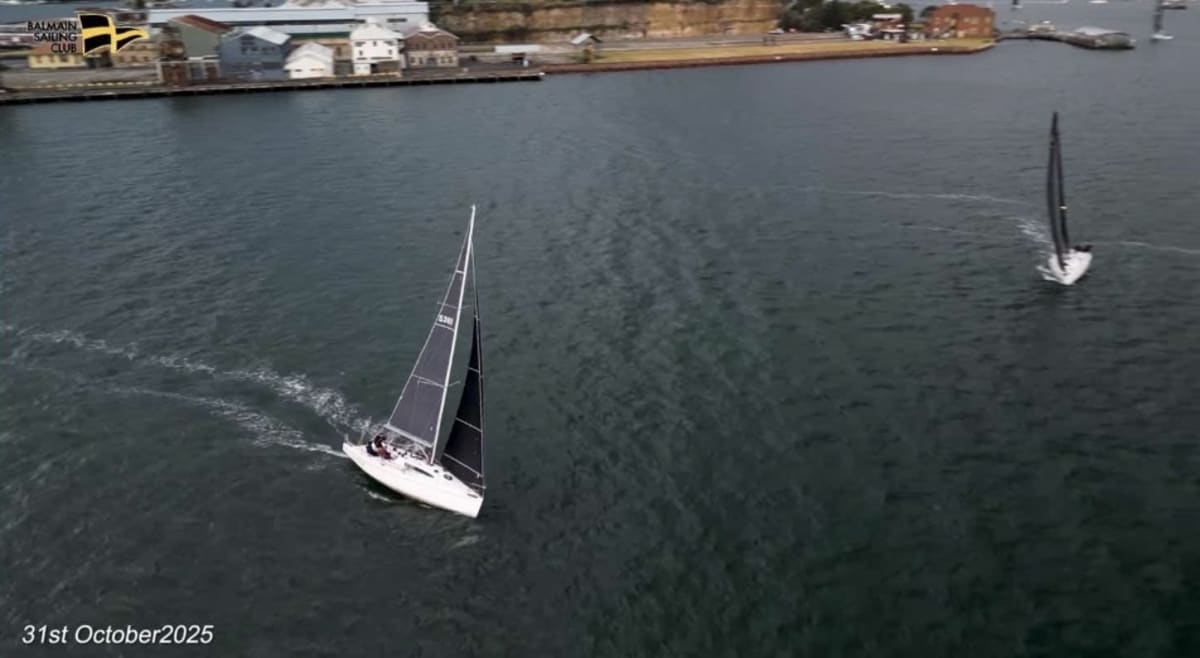 Two yachts racing on a quiet harbour near industrial shoreline on an overcast day.
