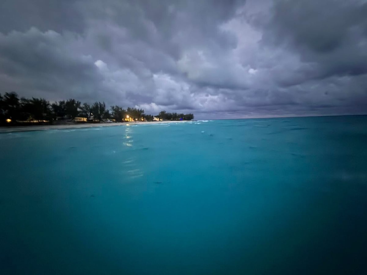 Calm turquoise waters under a moody sky at dusk near a shoreline with glowing lights.