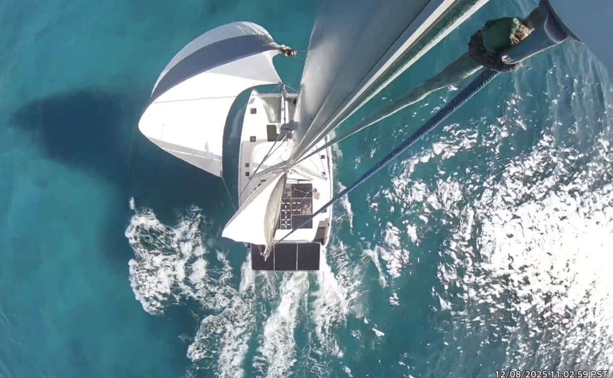Aerial shot of a catamaran sailing through clear turquoise water with sails full.
