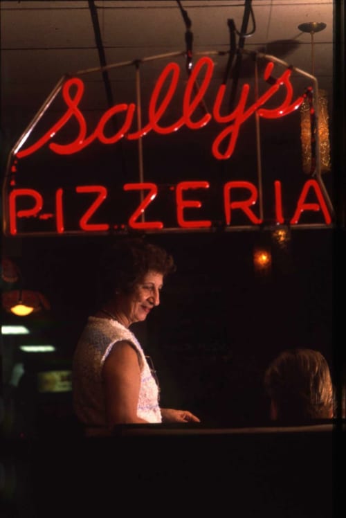 Flo standing under Sally's Apizza neon sign