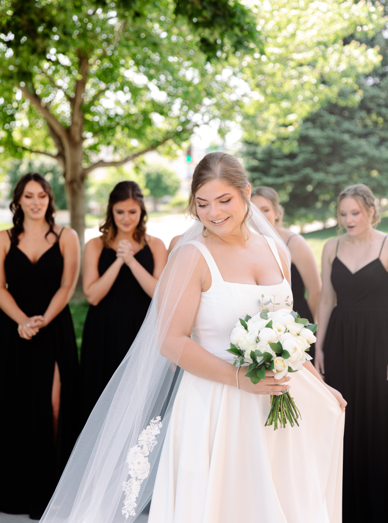 Bride holding flowers and wearing a wedding dress and a veil outside with her bridesmaids