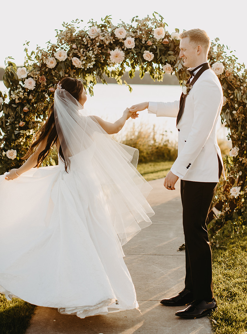 Bride and groom dancing outdoors in front of a flower arch with a veil twirling around the bride