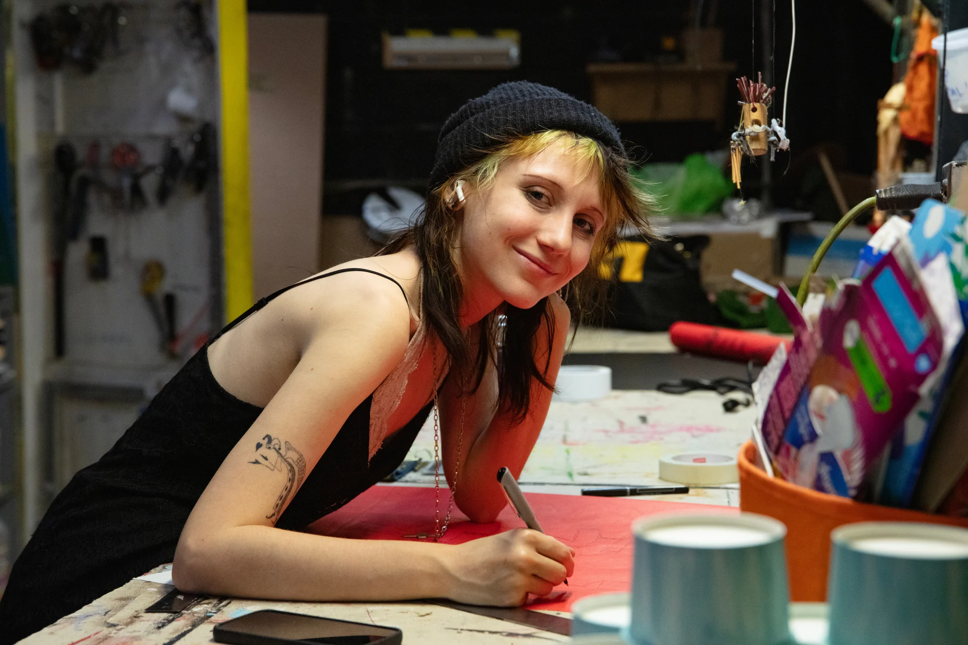 Person with short hair smiling, wearing a beanie and earbuds, leaning on a table while drawing with a marker. Art supplies are visible nearby.