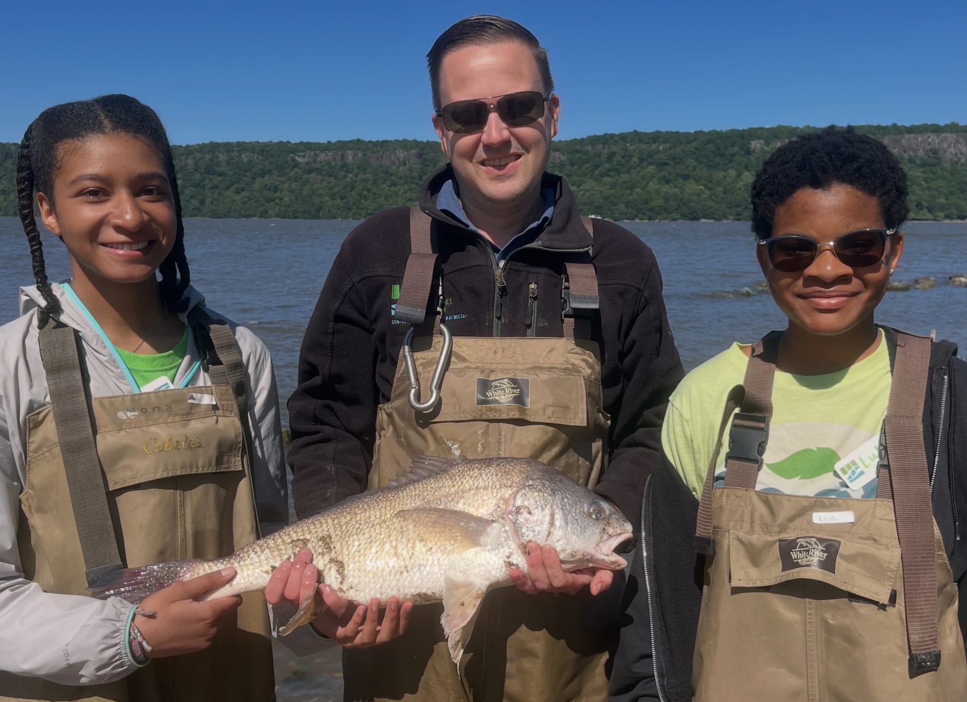 Three people in waders stand by a river, holding a large fish. The sky is clear with green hills in the background.