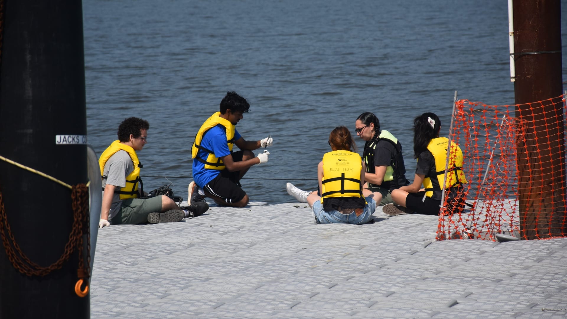 Five people wearing yellow life vests sit on a dock by a body of water, engaging in conversation or working with small objects.