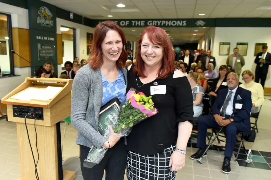 Two smiling women standing together, one holding a bouquet of flowers, with a crowd of people in the background and a podium to their left inside a building with a "Hall of Fame" sign.
