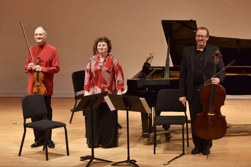 Three musicians standing on stage with a violin, cello, music stands, and a grand piano in the background. They appear ready for a performance.