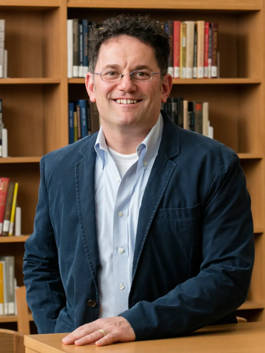 Person with glasses smiling, wearing a blue blazer and white shirt, standing in front of bookshelves.
