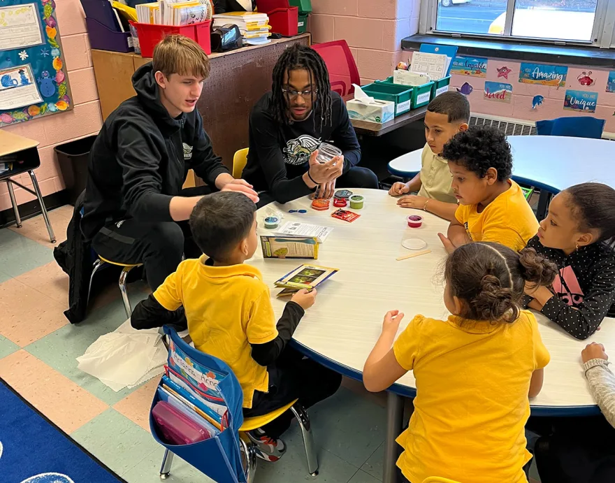 Two adults engaging with six children around a table with art supplies in a classroom setting.