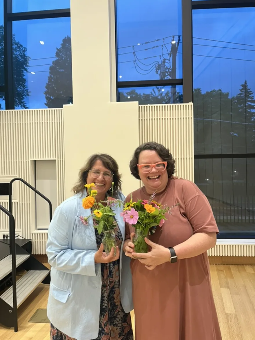 Two individuals smiling and holding vases with colorful flowers, standing in a well-lit room with large windows and a view of trees outside.