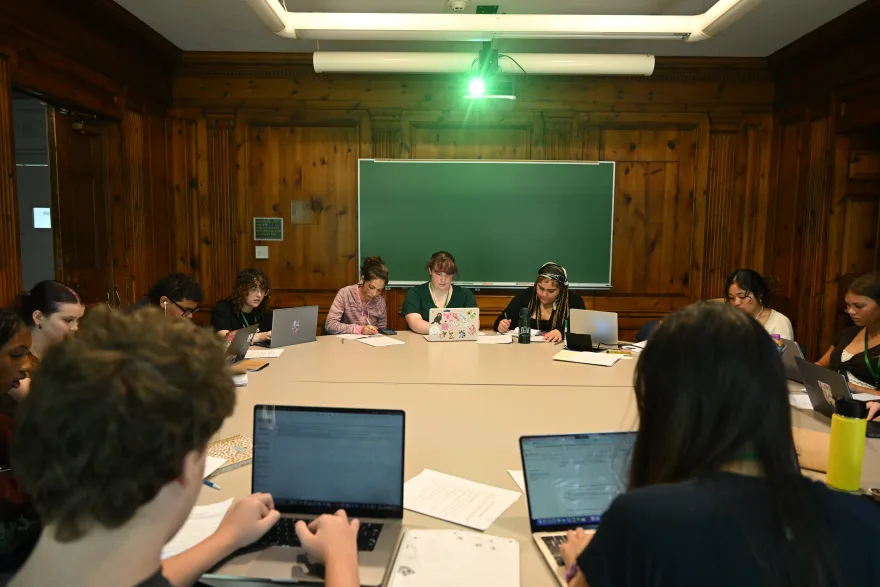 A group of people sit around a large table with laptops and papers in a wood-paneled room, under a lit projector.