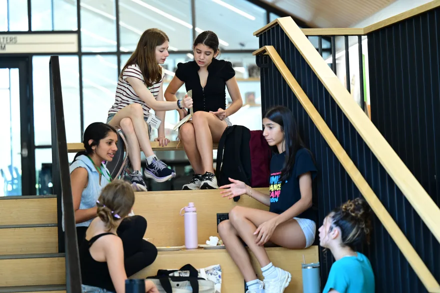 A group of young people sitting on indoor stairs, talking and holding notebooks. Backpacks and water bottles are nearby.