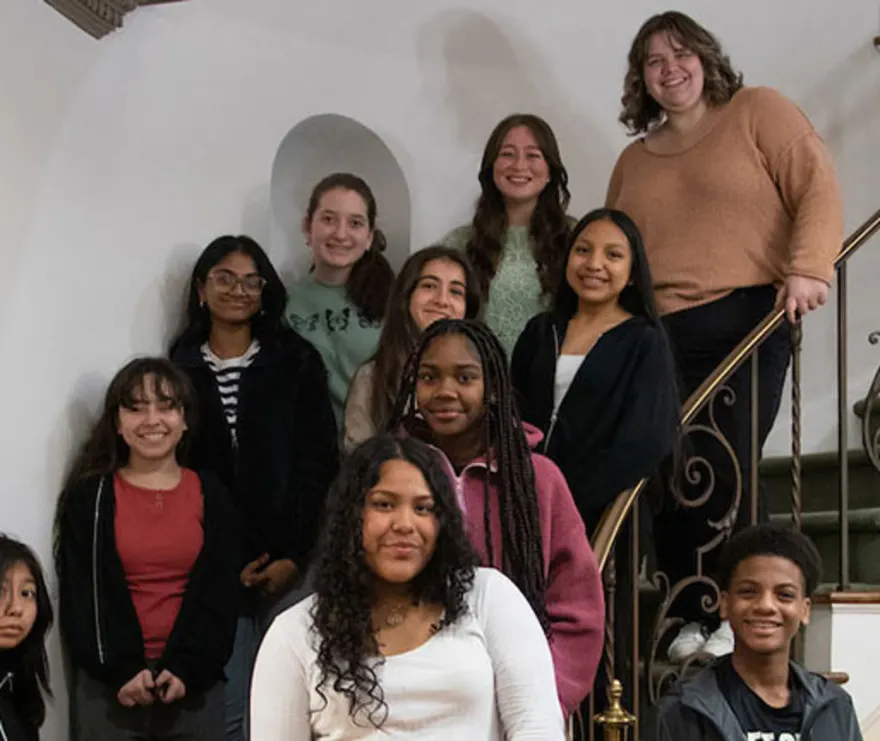A group of eleven people smiling on a staircase, with some standing and others sitting, in an interior setting with a white wall background.