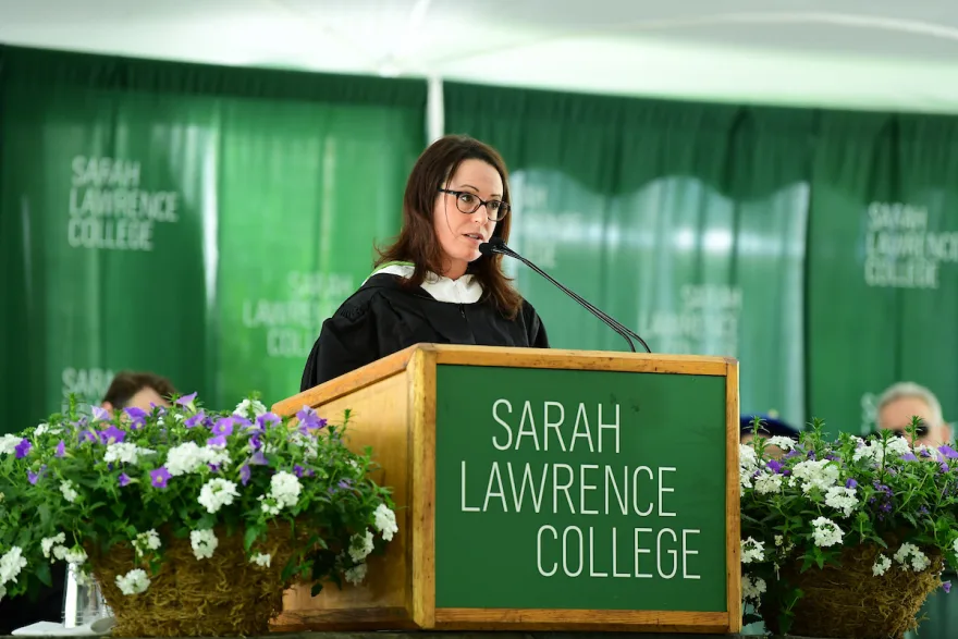 Speaker at podium with Sarah Lawrence College sign, surrounded by green curtains and floral arrangements.