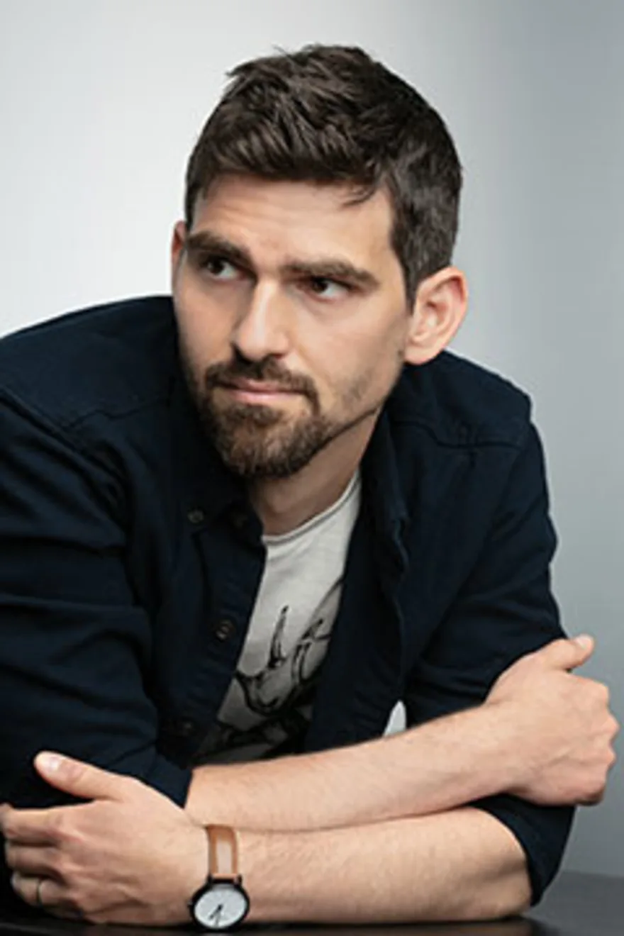 Person with short dark hair and a beard, wearing a black shirt and white t-shirt, looking to the side with arms crossed, against a gray background.