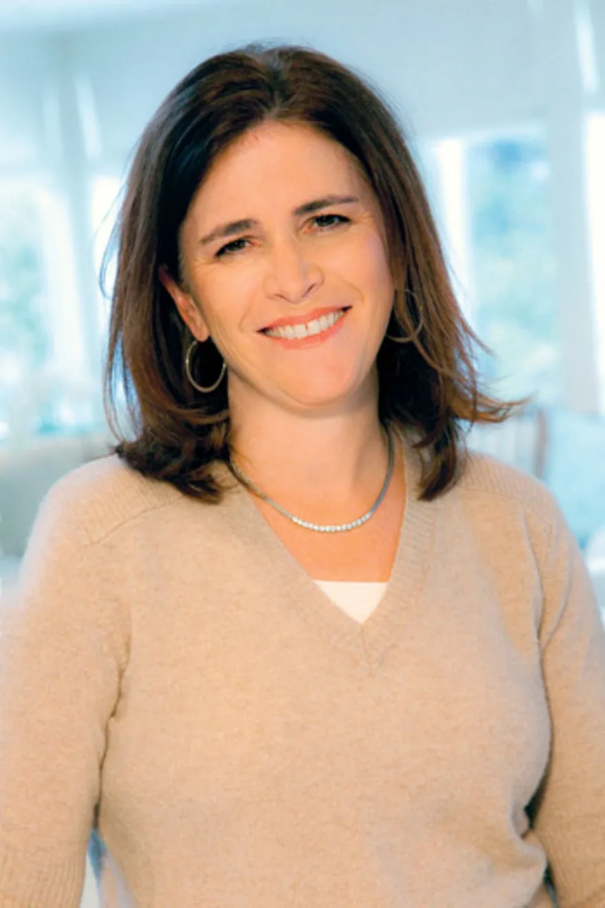 Person smiling, wearing a beige sweater and necklace, with shoulder-length brown hair, standing indoors with soft lighting.