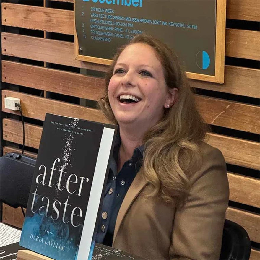 Person smiling while seated behind a table with a book titled "Aftertaste" by Daria Lavelle. A wooden wall and schedule board are in the background.