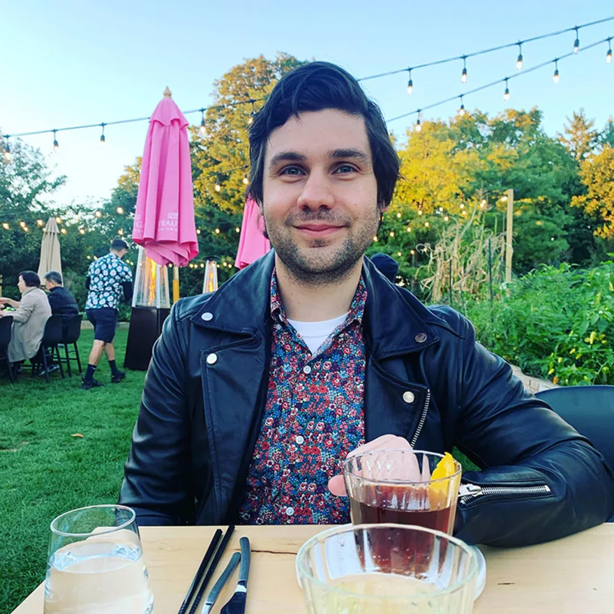 Person sitting at an outdoor table, smiling, with drinks and a leather jacket, string lights overhead, and trees in the background.