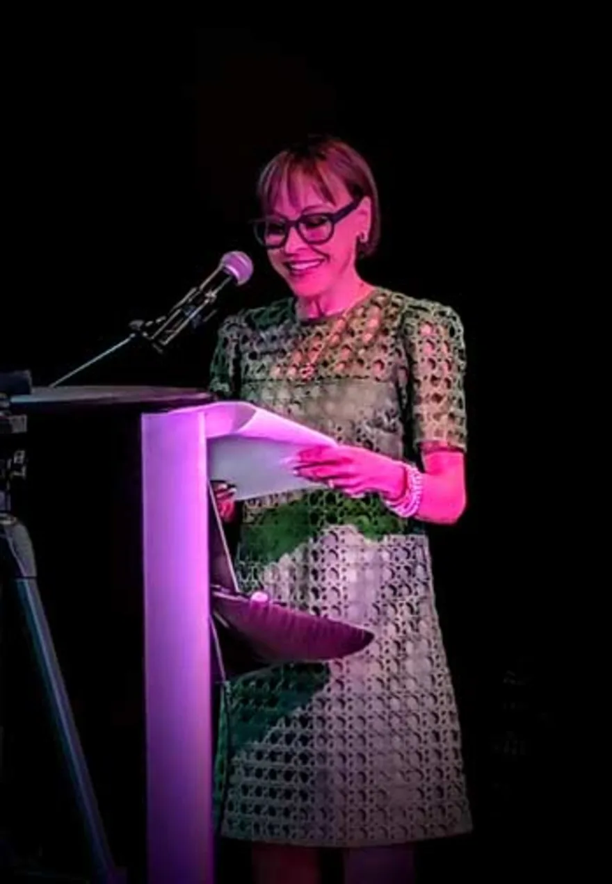 Person wearing glasses and a patterned outfit speaks at a podium with a microphone, holding a piece of paper, against a dark background.